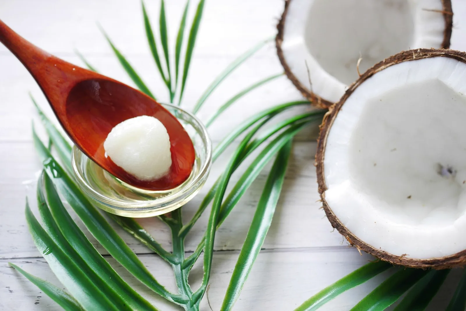 A wooden spoon of solid coconut oil over a glass dish, beside halved coconuts and palm leaves on white wood