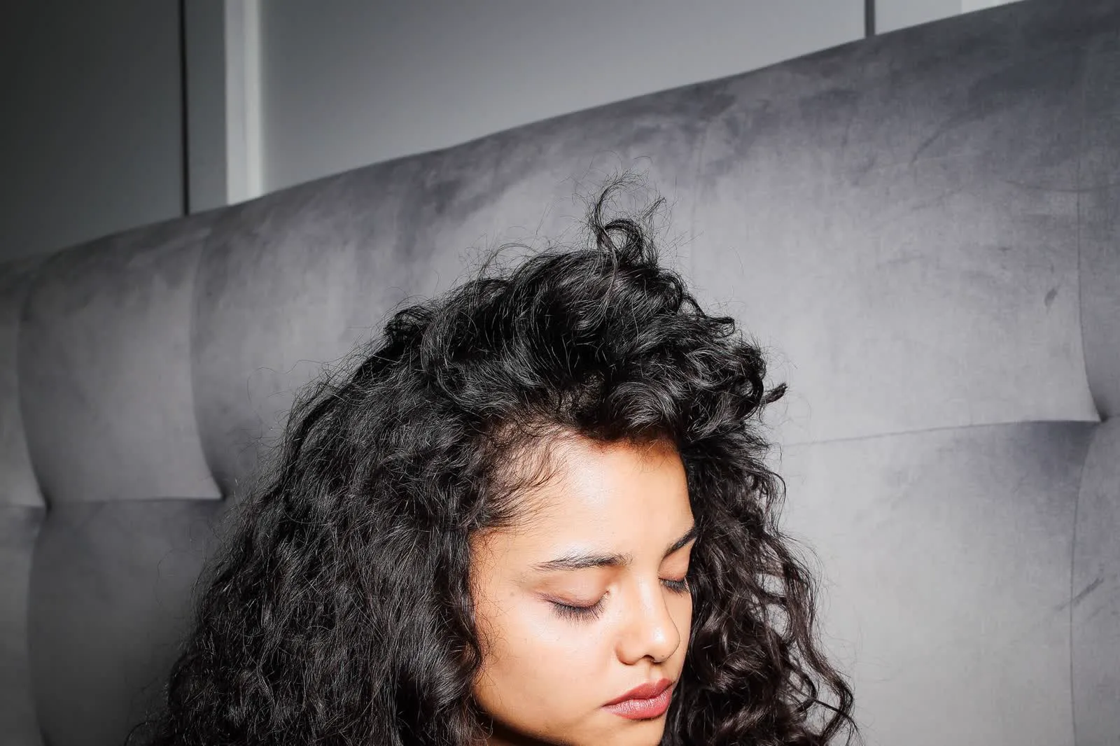 A woman with dark, defined type 3 curls resting against a soft grey headboard