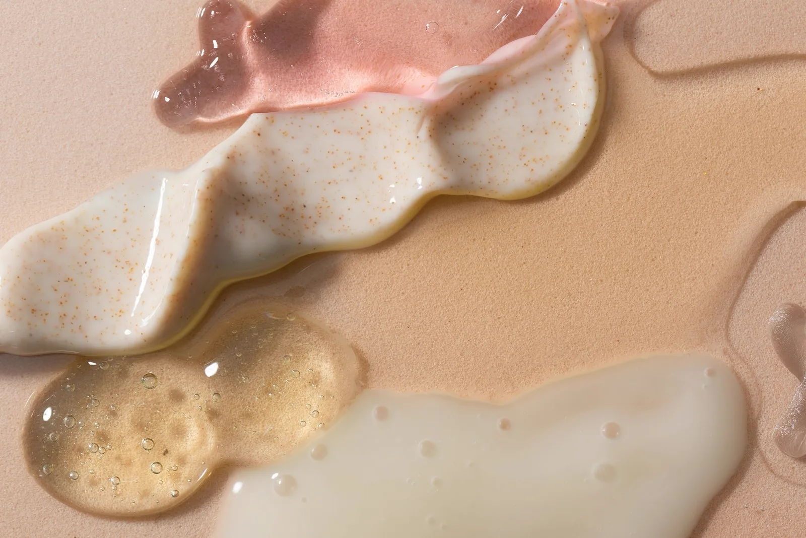 Smears of different translucent gel, cream and golden hair-product textures on a soft pink surface