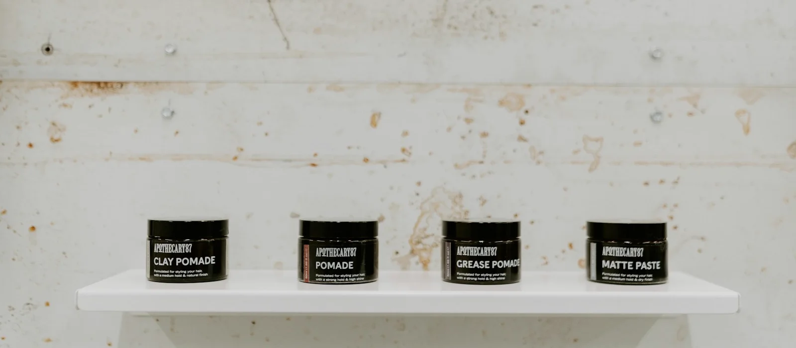 Four black jars of hair styling product lined up on a white shelf, labelled clay pomade, pomade, grease pomade and matte paste