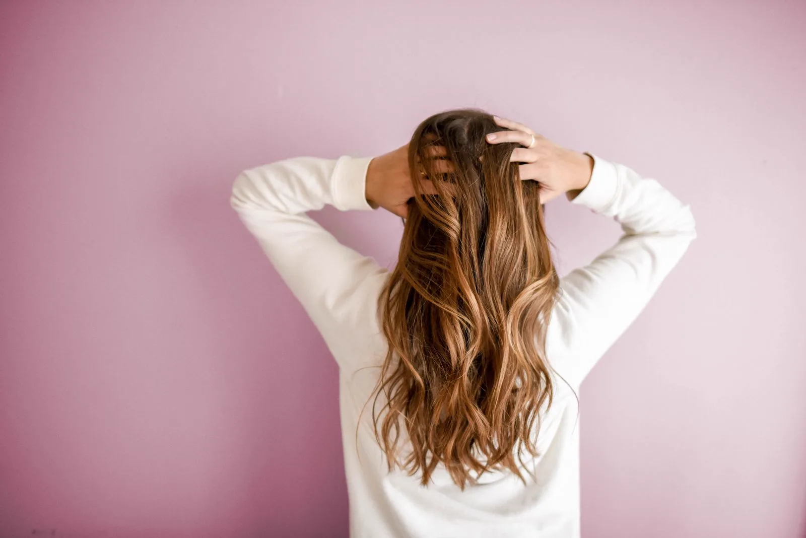 A back view of long wavy hair being lifted and scrunched with both hands against a pink background