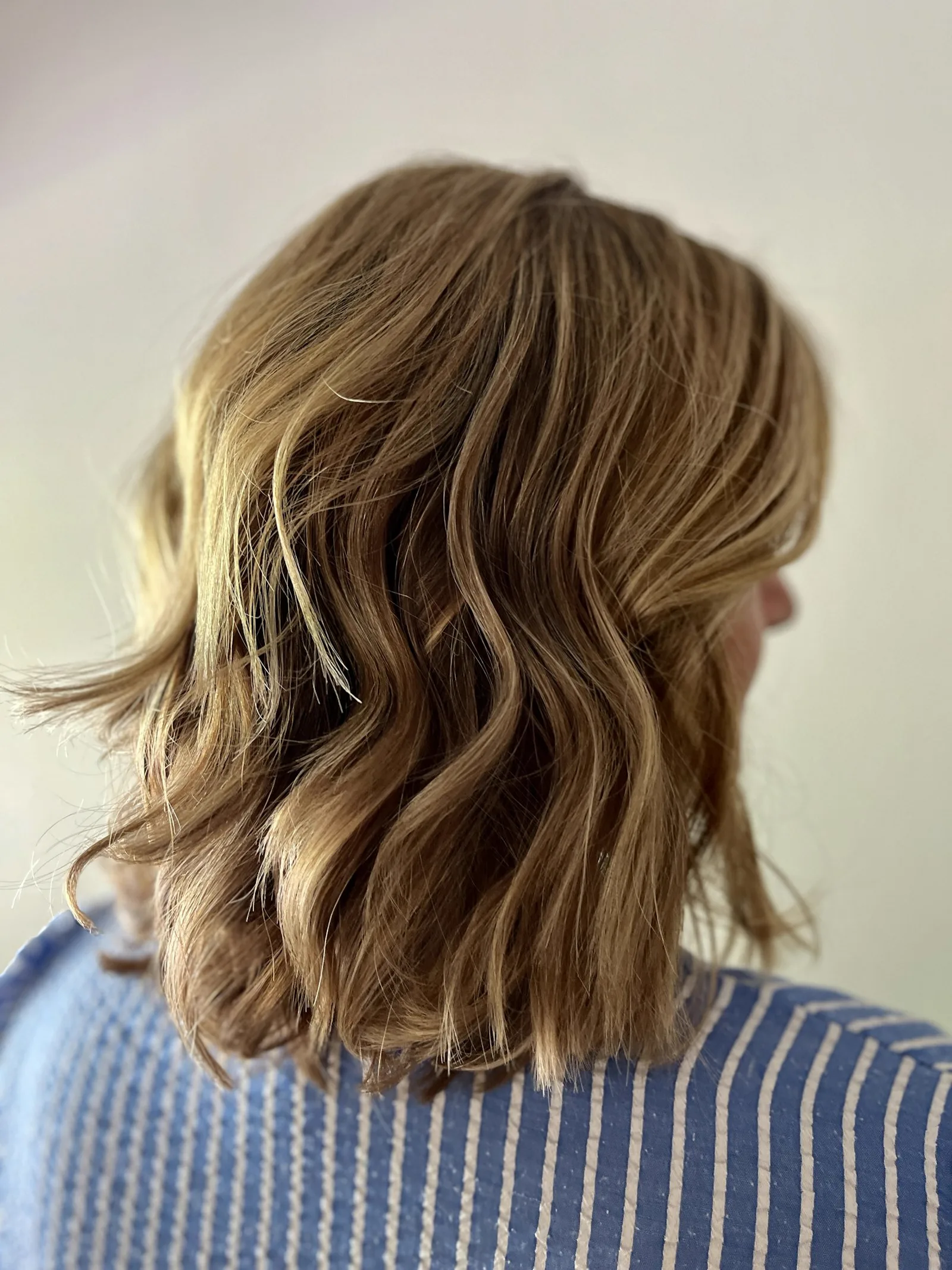 The back of a person's head showing shoulder-length light-brown hair with defined S-shaped waves
